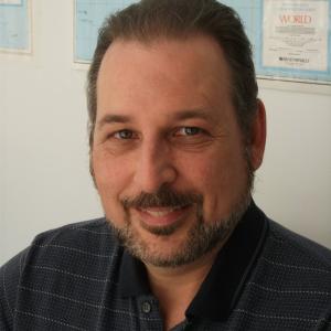 middle aged white man with a beard and mustache wearing a navy blue shirt sitting in front of a world map