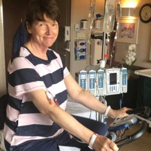 middle aged white woman with short brown hair wearing a navy and pink striped blouse and blue pants riding a stationary bike in a hospital room