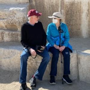 older white couple sitting in front of stones man wearing burgundy ball cap and black shirt and woman wearing blue shirt face maske and straw hat