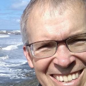 older white man with brown framed glasses plaid shirt and black jacket and big smile standing in front of the ocean