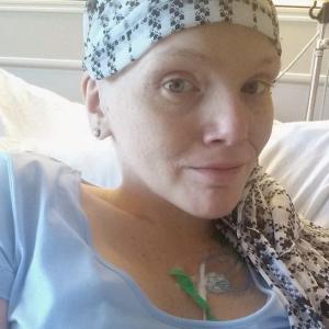 young white woman with a scarf on her bald head wearing a blue shirt laying in a hospital bed