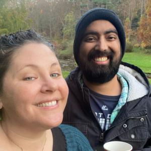 young white woman in blue sweater holding coffee with young black man with beard and mustache wearing a knit cap and blue jacket holding a cup
