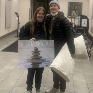 Flavio young hispanic couple she is holding Team Flavio poster and he is holding a pillow both wearing black jackets