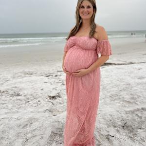 Holly young white woman with long hair and very pregnant wearing a pink off the should lace dress standing on a beach