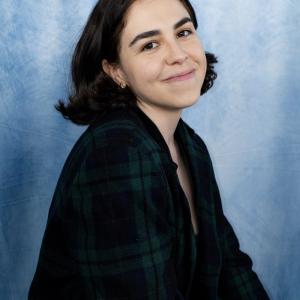 Crystal young hispanic woman with dark hair and gold hoop earrings smiling and wearing a blue and plaid top with blue photo background