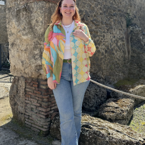 Eva young white woman with reddish hair sunglasses on her head in a white tshirt under a multi color shirt with blue jeans standing in front of a stone wall.