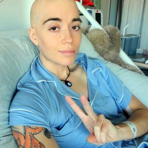 Josie young white woman bald with an orange tatoo and wearing blue pajamas in a hospital bed giving the peace sign