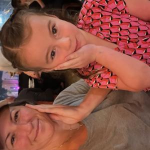 Kacy young white woman wearing ball cap and greenish tshirt with daughter in a pink and black top and both have a hand up against their cheeks