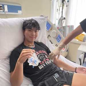 Kevin young asian young man with black hair wearing a puka shell necklace and black gym tshirt and shorts while laying in a hospital bed