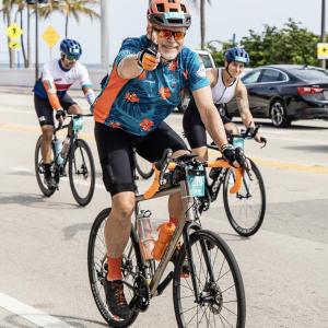 Michael older white man with white beard and mustache wearing an orange bike helmet and blue and orange shirt riding a bike while giving a thumbs up