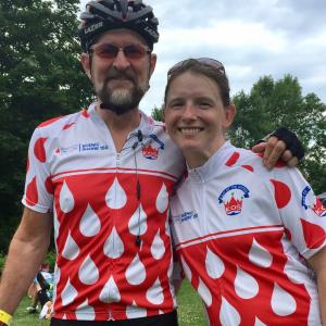 Paul older white gentleman with beard and mustache wearing a bike helmet and sunglasses with younger white female with freckles and sunglasses on her head both wearing Scenic Shore 150 shirts