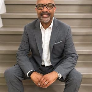 Arturo Pierre middle aged black man bald with mustache and beard wearing black glasses and a grey suite and white shirt sitting on steps