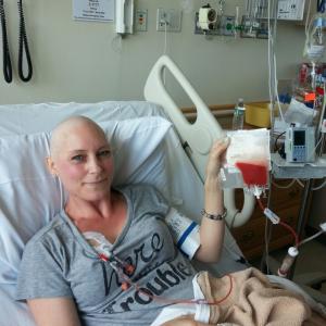 Babette young white woman who is bald waring a grey t-shirt and lying in a hospital bed holding an infusion bag