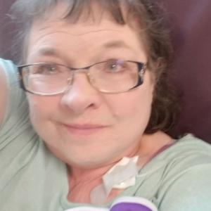 Dixie older white woman with brown hair and glasses wearing a green shirt laying against a brown pillow with an earbud laying on her chest