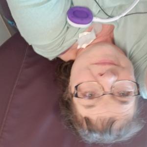 Dixie older white woman with brown hair and glasses wearing a green shirt laying against a brown pillow with an earbud laying on her chest