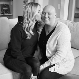 Janie young white woman who is bald wearing a black shirt and a white speckled cardigan being kissed by a young white woman with long blond hair wearing a black hoodie both sitting on a couch