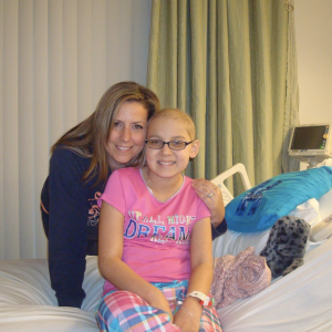 Kendra woman and young hispanic girl on a hospital bed girl has glasses and wearing a pink shirt and plaid pajama bottoms with turqouise pillow on bed