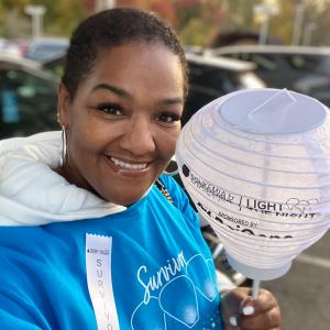 Kenya middle aged black woman with short black hair wearing big hoop earrings and a blue survivor tshirt and white ribbon hold a LTN light