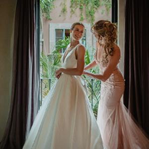 Laura young white woman in a white wedding dressing looking at her mother who has a curly hairdo and wearing a pink formal dress both standing in front of a balcony