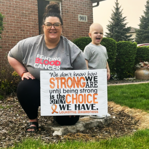 Maverick young white mother with a bun and glasses wearing gray blood cancer shirt holding a leukemia sign with her young son next to her in a gray shirt and smiling
