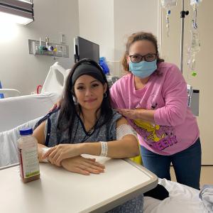 Mayra young hispanci woman sitting in a hospital bed wearing a black headbank and blue hospital gown and has three face piercings with another woman with face mask and pink shirt next to her