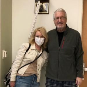 Rex older white couple standing in a hospital room man has white hair and glasses wearing a gray jacket and pajama pants woman has face mask