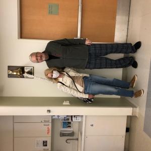 Rex older white couple standing in a hospital room man has white hair and glasses wearing a gray jacket and pajama pants woman has face mask