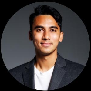 Rohan young man of Indian heritage with dark hair and slight chin stubble wearing a dark gray jacket over a white tshirt.