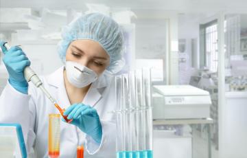 Researcher wearing a hair net, N95 mask and gloves injects a liquid into a vial.