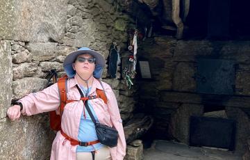 Kendra stands against a stone wall, wearing a pink shirt, hiking gear, and a hat.