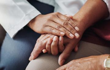 A person with dark-skinned hands clasps the hand of a second dark-skinned person to offer comfort.
