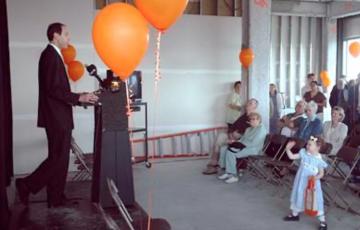 A man stands at a speaking podium, surrounded by orange balloons. He's facing a small crowd of people sitting in folding chairs or standing as they listen to him. A young girl wearing a dress and hair bow waves in the foreground.