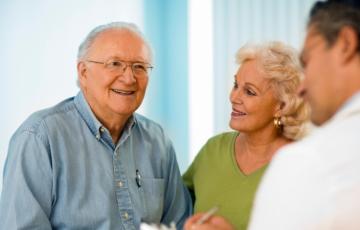 An older, white man smiles and is joined by his wife, who is also white, as he speaks to his doctor