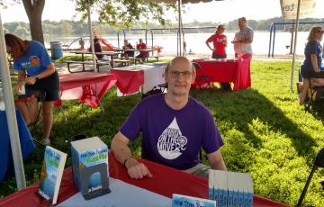 Steve, a white man with glasses, sits at a table with a stack of books. The table is surrounded by several other tables in a park. 