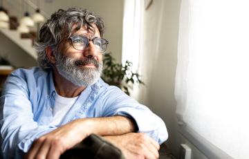 A content-looking man with long gray hair and a thick beard sits at home and looks out the window