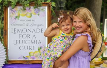 Blond girl in purple dress holding younger girl by lemonade stand sign.