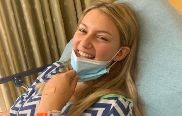 Blonde teen girl with leukemia in blue and white medical gown pulling down a face mask