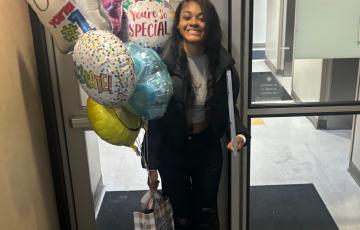 Smiling black teen girl with lymphoma in black jacket and pants holding mylar balloons and a gift bag