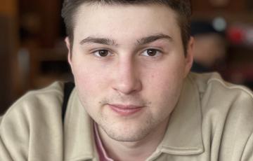 young white man with dark brown hair and chin stubble wearing a pink shirt and beige jacket