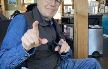 Older white man wearing glasses a denim shirt with a black vest and jeans pointing a finger.jpg