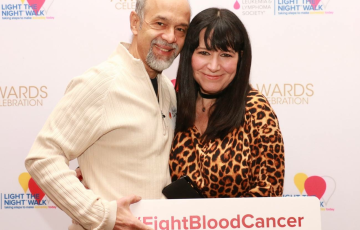an older hispanic couple hugging in front of LTN wall he in a cream shirt holding a sign that says FightBloodCancer and her in an animal print top