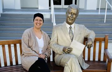 bald white woman wearing a knit cap tan sweater and white shirt sitting on a bench next to a statue
