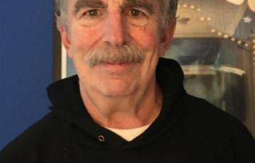 older white man with bushy eyebrows and a mustache wearing a black hoodie that says Italia standing in front of a movie poster