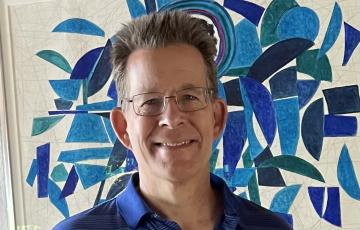 older white man with spiky hair wearing glasses and a blue striped shirt in front of mosaic artwork