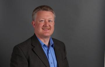 smiling middle aged white man with graying hair and mustache wearing a brown jacket and blue shirt