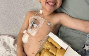 smiling young white boy laying in bed with a port in with a box of rolls