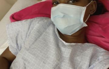 young black woman wearing a mask and hospital gown lying on a bed with a pink blanket