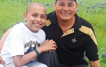 young hispanic child with little hair in white t-shirt and sweat pants with his father who is wearing a New Orleans Saints hat and a black and gold shirt, both in a field of bluebonnets