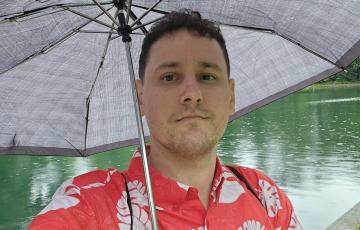 young man with scruffy beard and mustache in an orange and white shirt holding an umbrella in front of water