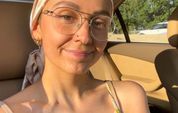 young white woman sitting in a car wearing a head scarf, wire rim glasses, a floral sundress witha nose ring
